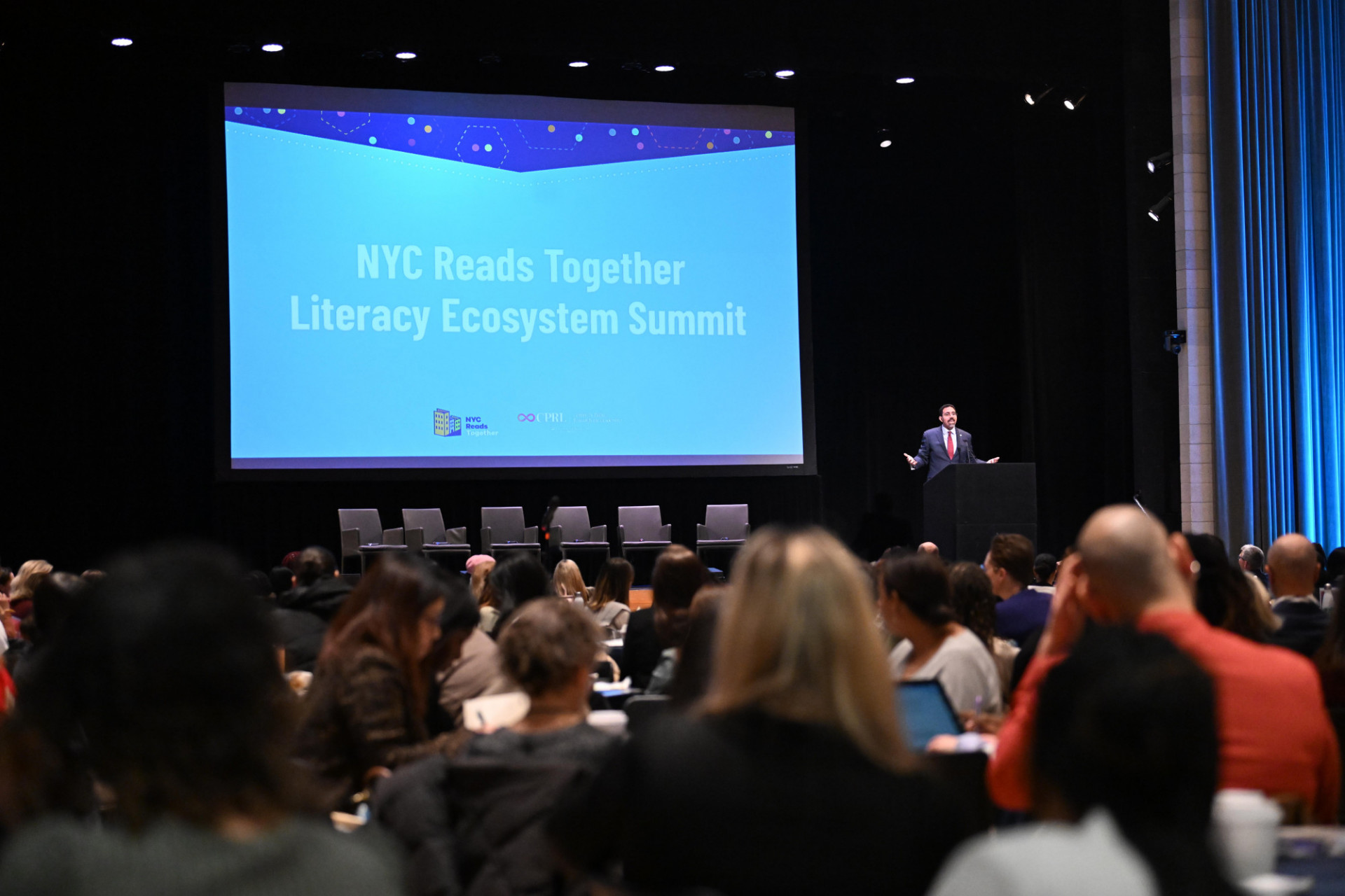 a screen with 'nyc reads together literacy ecosystem summit' and a speaker talking to an auditorium of people