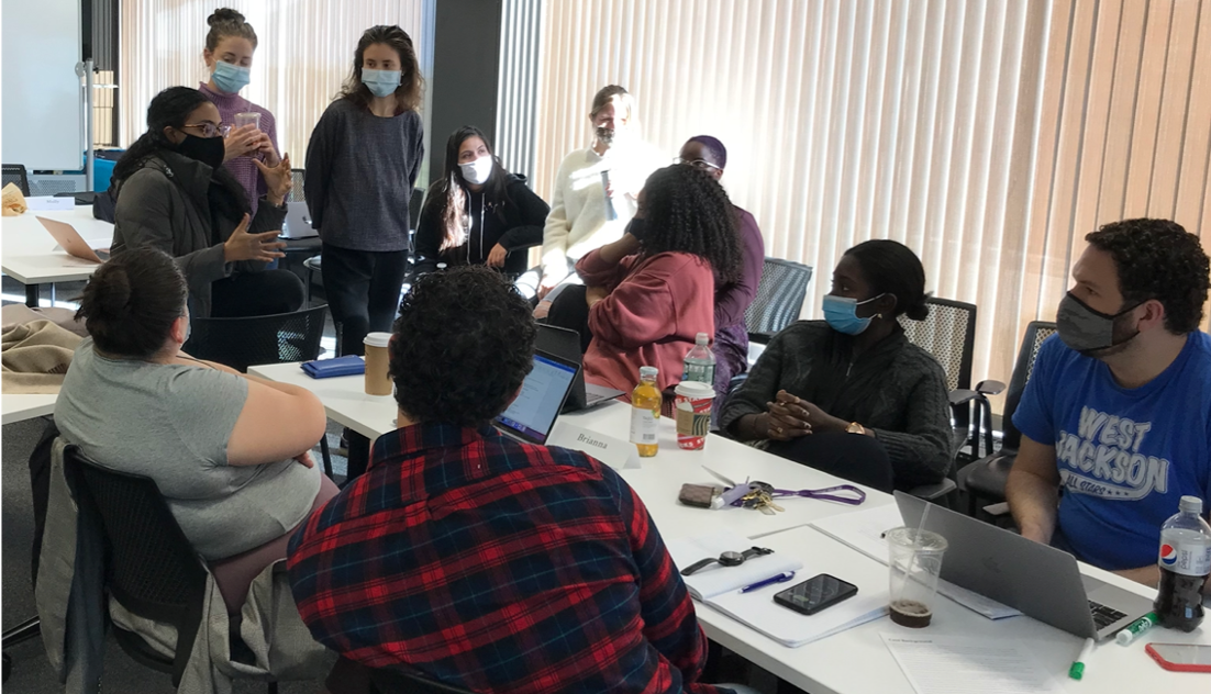 CPRL students seated and standing around a table talking with each other.