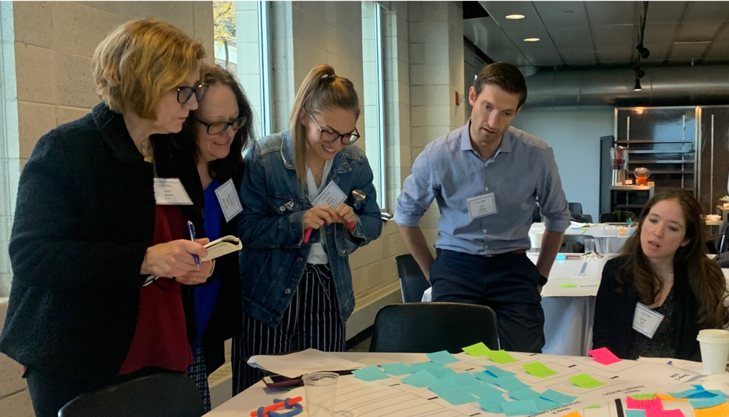 Five people seated and standing around a table looking at a chart