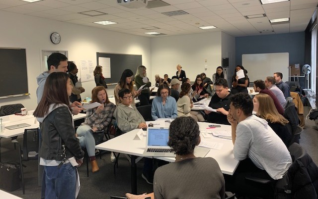 Approximately 30 graduate students seated and standing around a table talking and listening