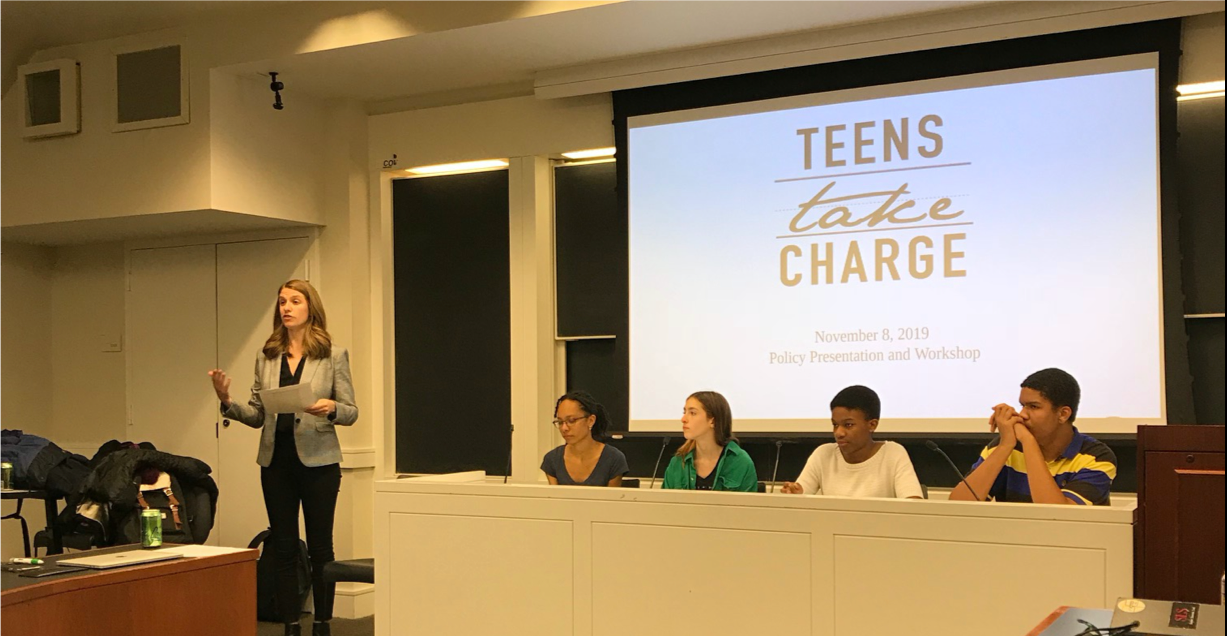 A discussant standing alongside a panel of four young adults who are seated; The image "Teens Take Charge" appears behind them on a screen.